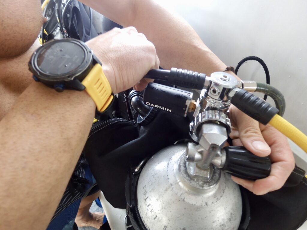 Garmin Descent dive computer on wrist next to scuba diving tank with garmin descent wireless air integration transmitter.