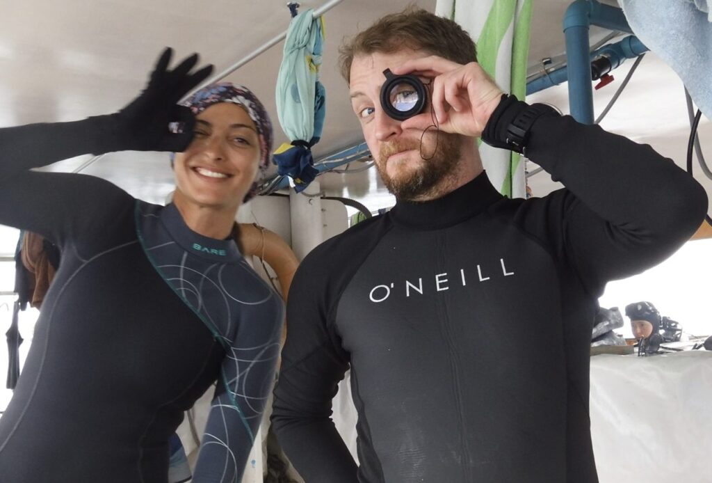 Two divers (Alex and Tsvetelina) standing on a Donny in wetsuits giving the diver OK sign.