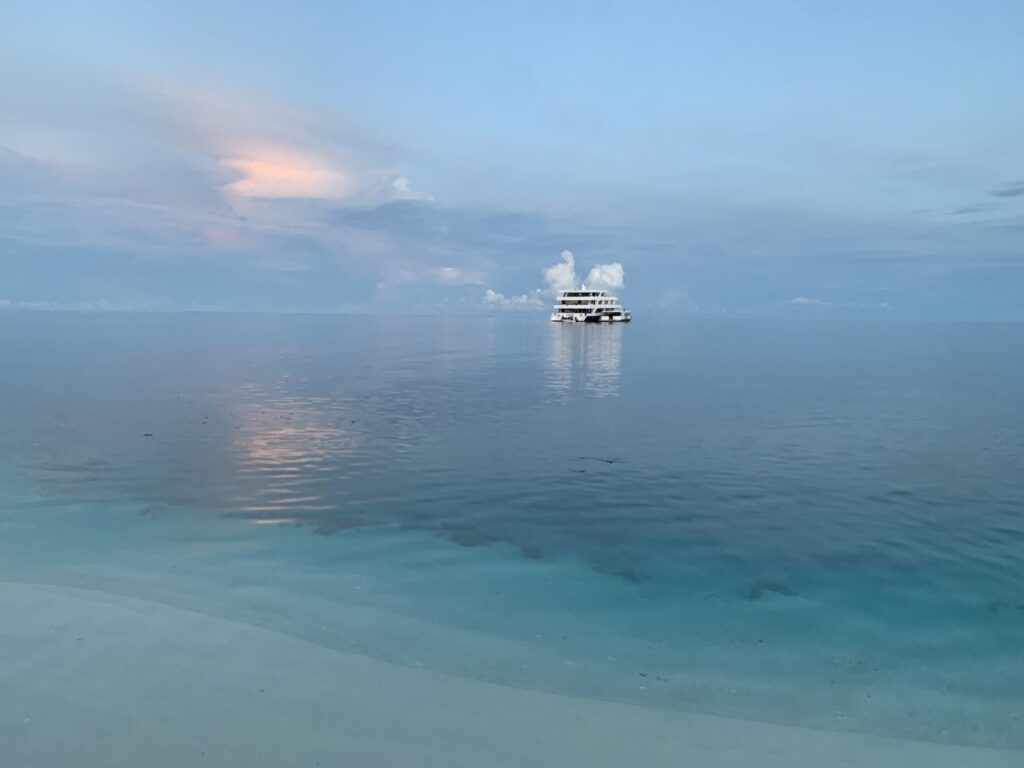 Maldives liveaboard on ocean in distance against pale blue sky with sunset.